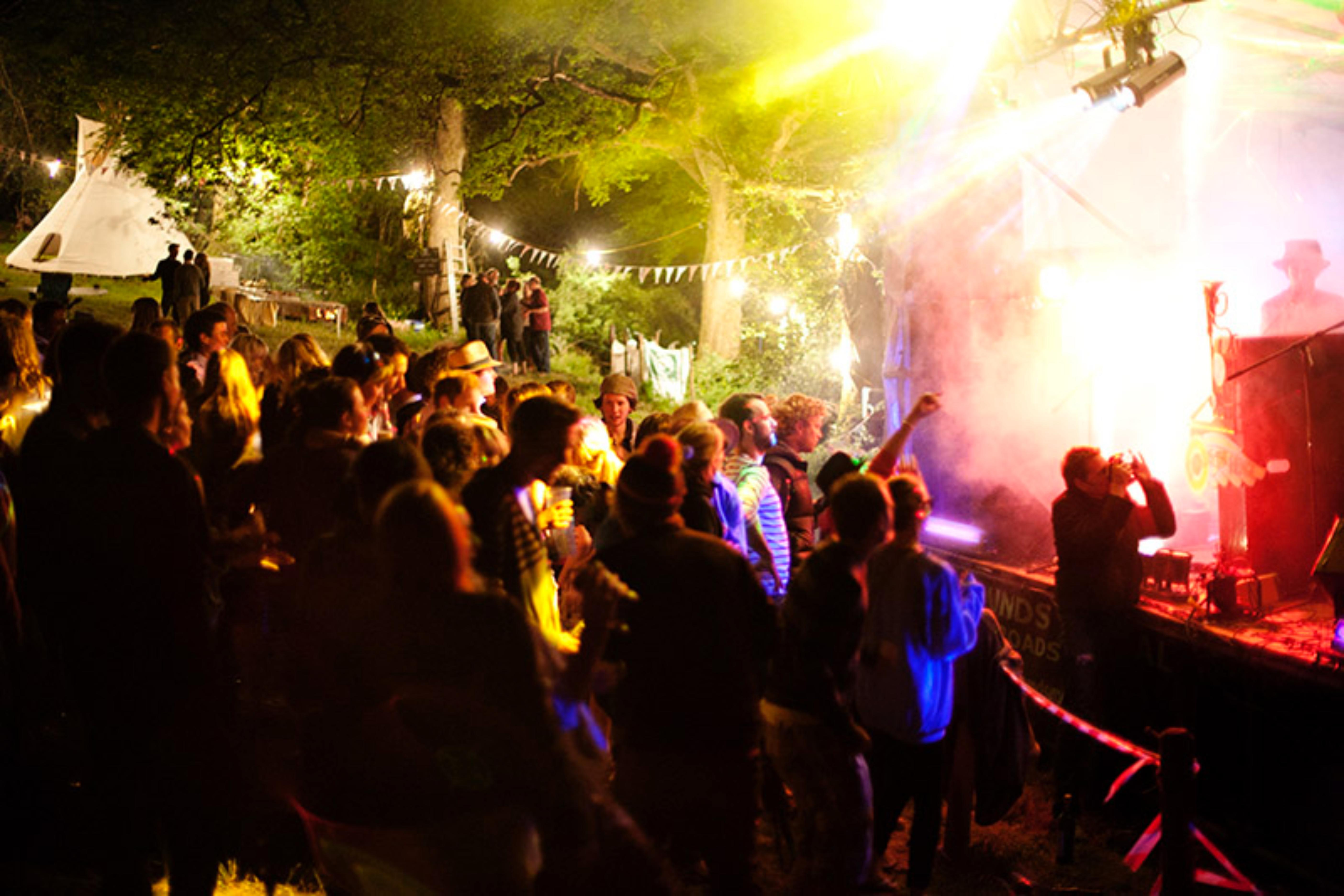 People watching a band at a woodland festival, A small crowd enjoying a band playing on the stage in front of them. It is night time and the stage is lit by colourful lights. They are in a woodland festival setting with bunting and tents in the background.