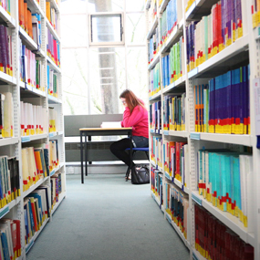 A corridor of library shelves with a woman sat at a table at the end 