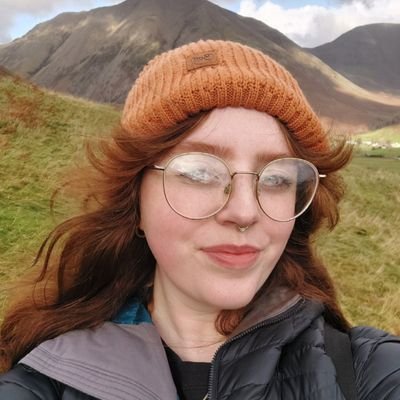 A lady standing before a mountain with red hair and orange hat and glasses