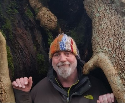 Head shot of man with beard in a wool beanie hat standing in front of a large tree