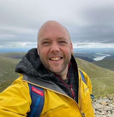 A head and shoulders selfie image of a man wearing a yellow waterproof jacket standing with a mountain landscape behind him