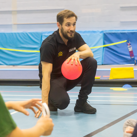 A male in black clothing kneeling down to the floor in a sports hall. His right hand is touching the floor, while his left is holding onto a pink ball.