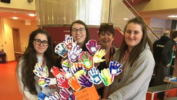 WCF Students, Four females stand together holding an art collage of painted, colourful hands, and smiling at the camera.