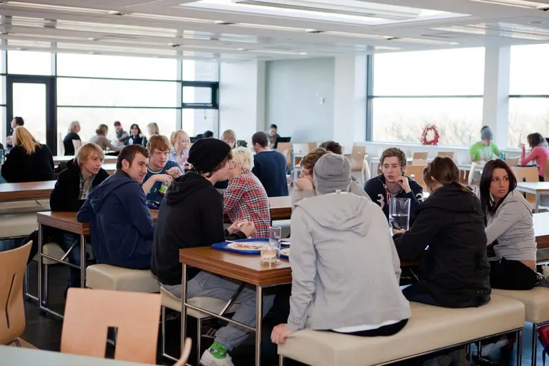 A large sunlit inside dining area with several long tables full of seated students.