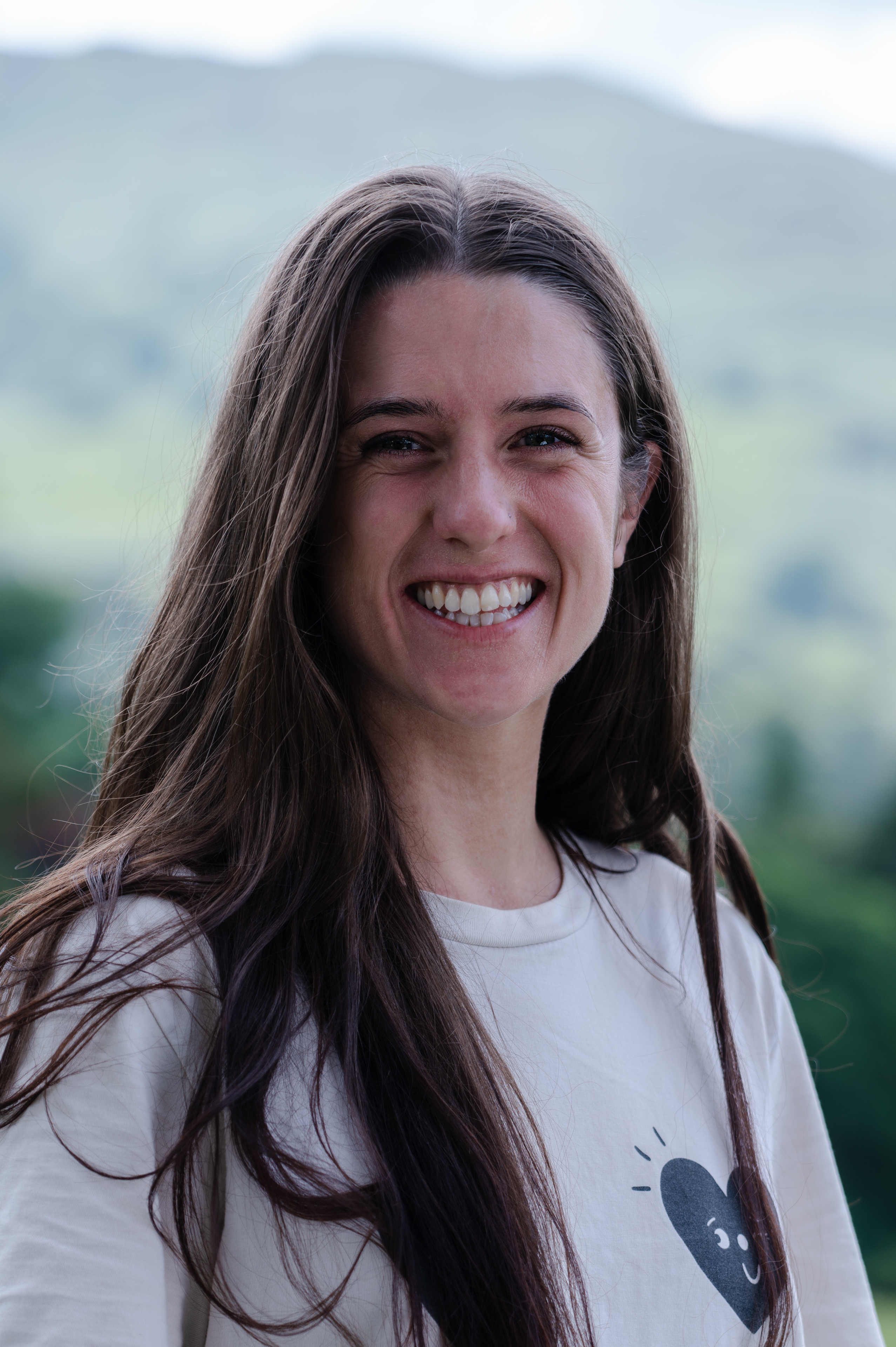 Molly McConnell New, Portrait of a long dark haired female smiling at the camera. She is wearing a white top with a heart logo on the top left.