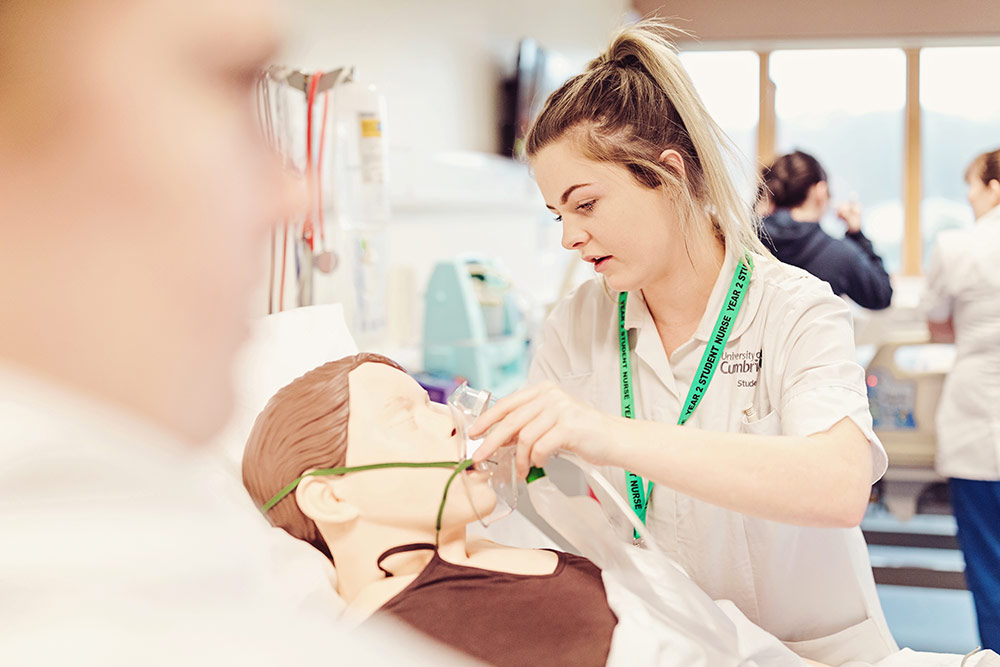 Nurse , A female student wearing her university tunic applies an oxygen mask over the face of a patient mannequin. 