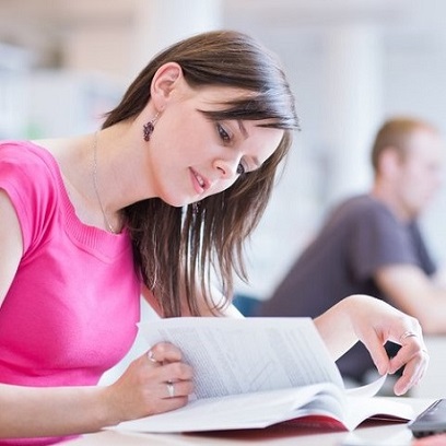 A female in a pink top sits at a desk and flips through a textbook.