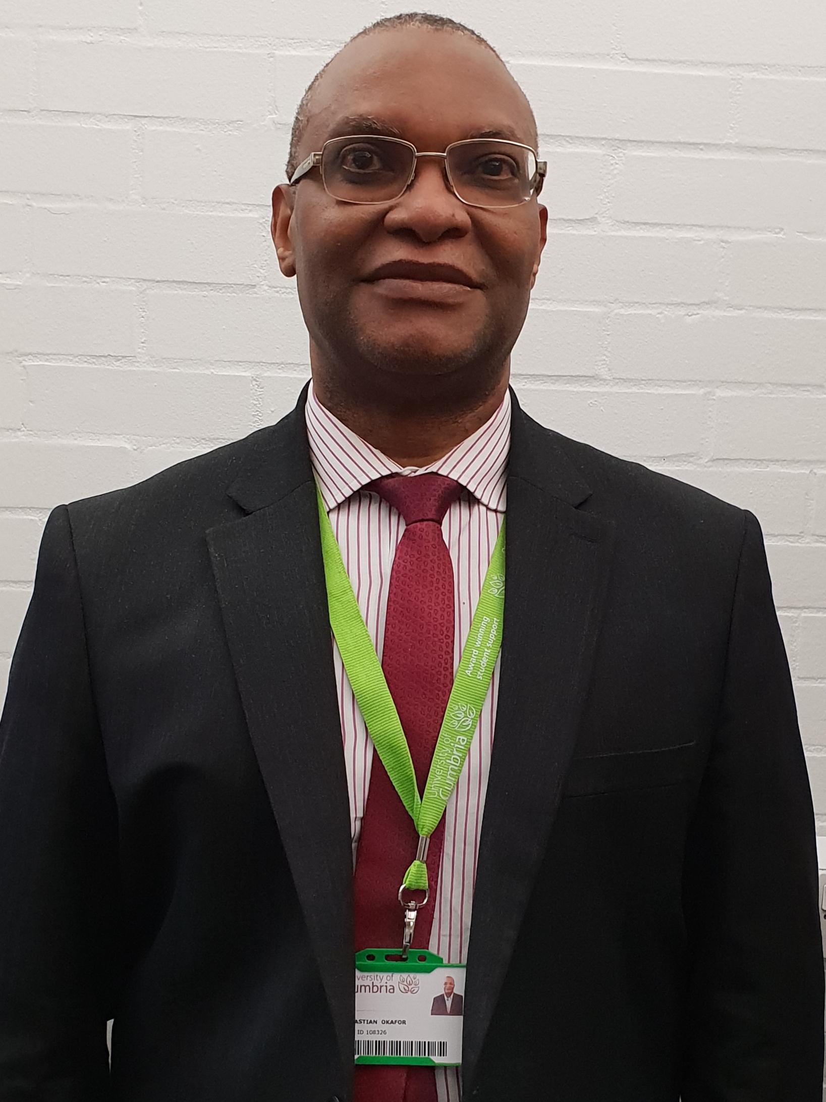 Portrait of a man wearing a smart black suit, red tie and glasses. A university lanyard is round his neck.