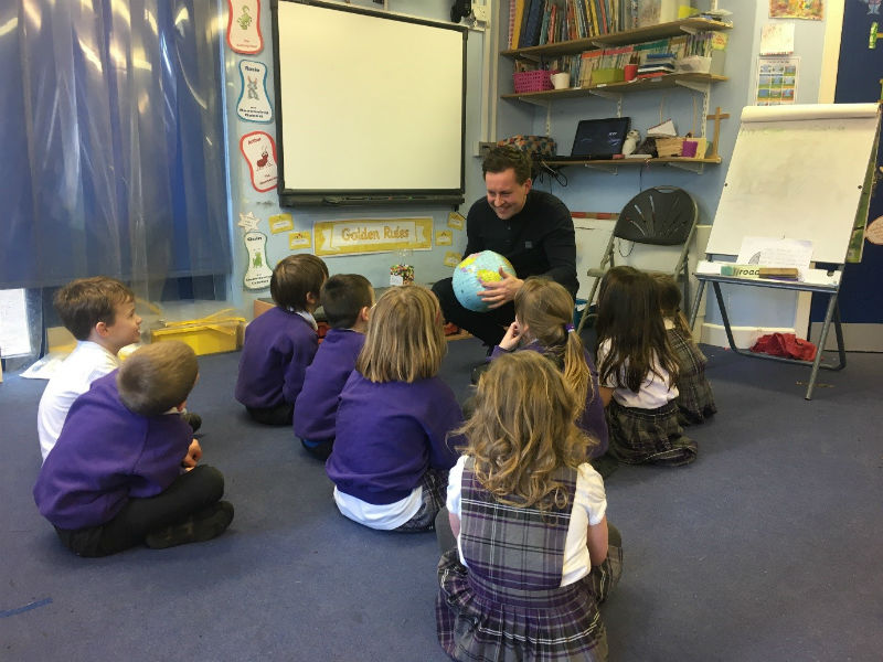 Steafan Deery during his placement in north Lancashire, A classroom setting. A man crouches down to the ground while holding a globe in his hands. School children sit on the ground around him.