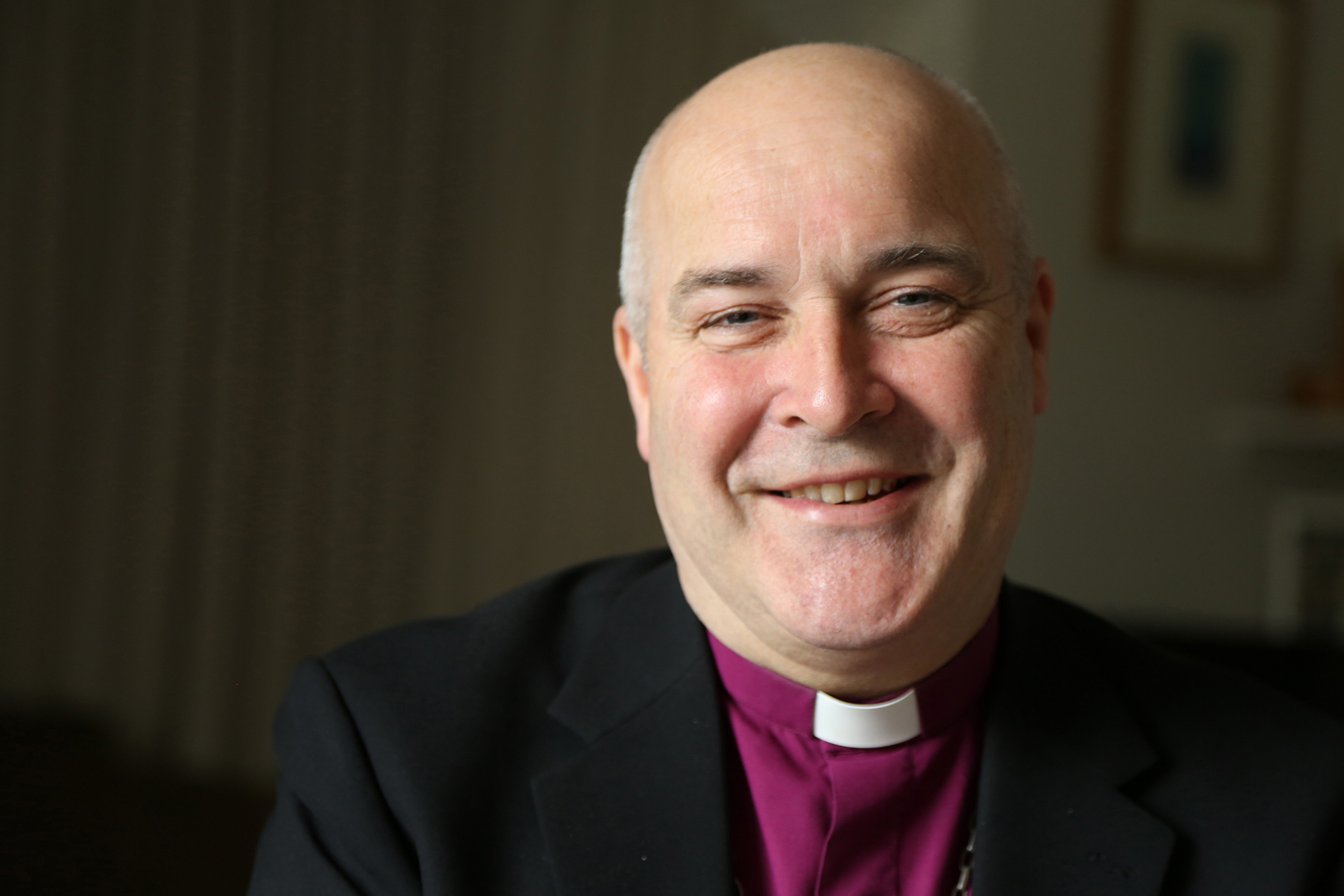 Picture of a vicar  dressed in a black suit and purple shirt smiling at the camera.