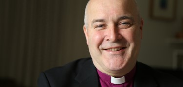 Picture of a vicar dressed in a black suit and purple shirt smiling at the camera.