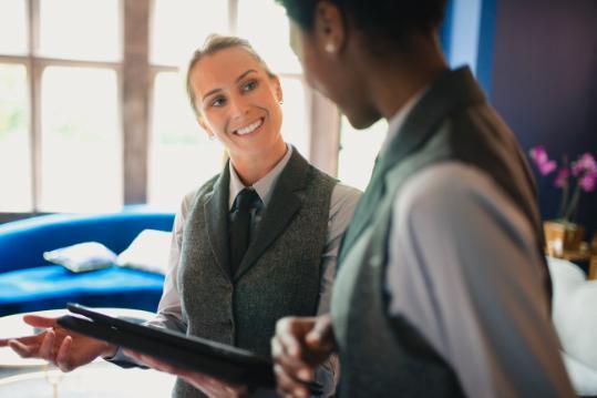 13_reasons_app, Two smartly dressed females, one holding an electronic device, are smiling and conversing with one another.