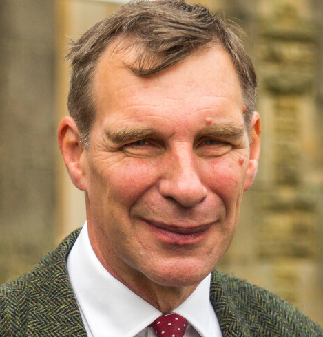 A man in a white shirt and tweed jacket smiles at the camera.