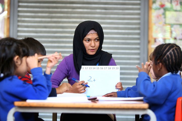 Female Primary School Teacher, female primary school teacher, sat down with 4 young children, holding a small whiteboard