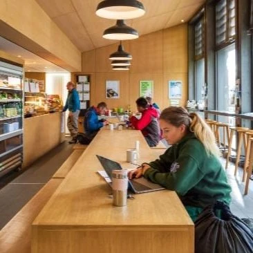 Several students eating and chatting at the far end of a long bench in a modern canteen.
