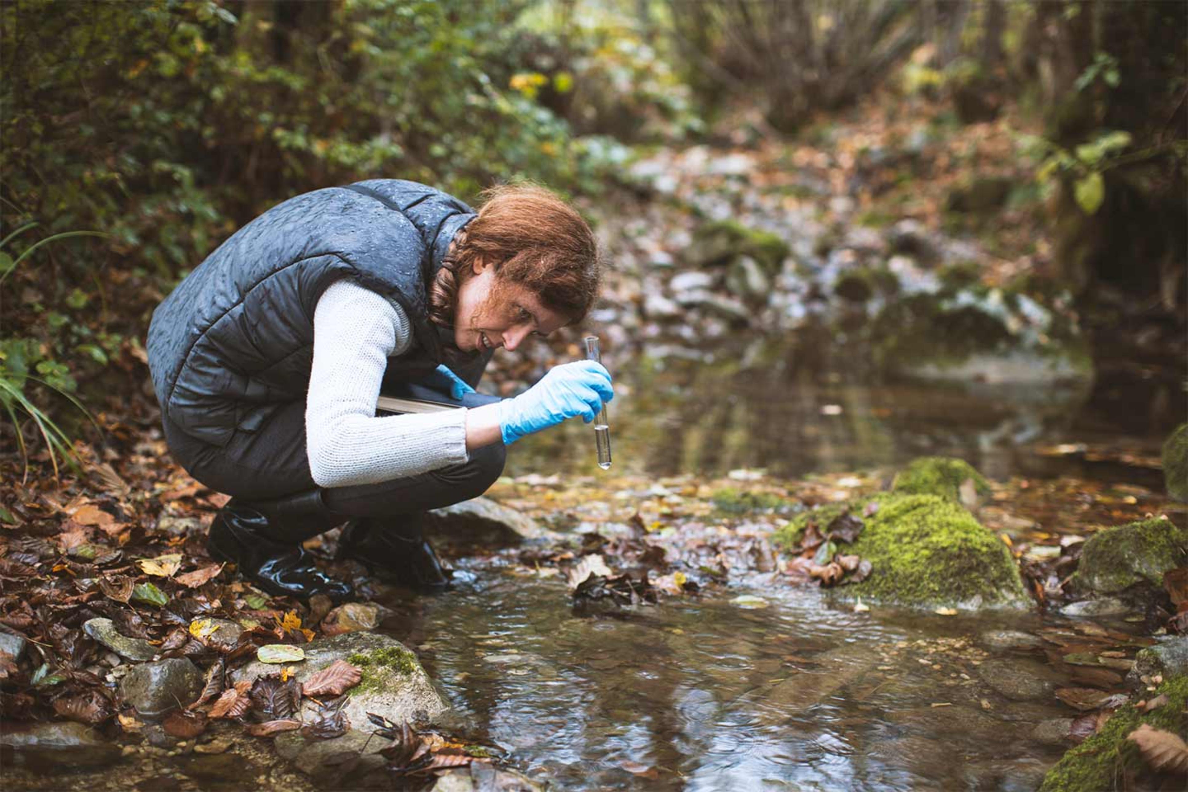 A woman sampling water from a stream , A woman crouching down by a stream and inspecting water she has collected in a test tube. 