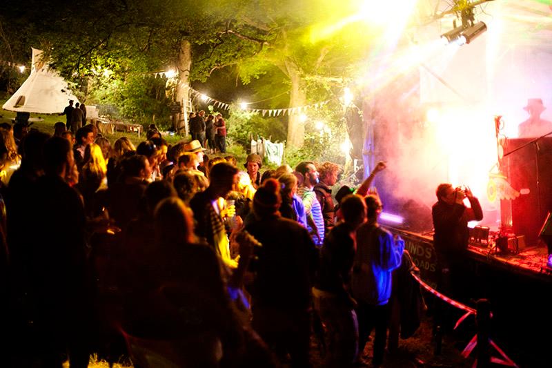 People watching a band at a woodland festival, A small crowd enjoying a band playing on the stage in front of them. It is night time and the stage is lit by colourful lights. They are in a woodland festival setting with bunting and tents in the background.