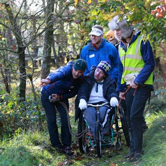 A photo of a wheelchair user being assisted through a wooded area by other students/staff