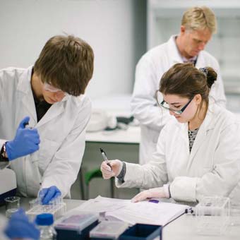A photo of two students in lab coats conducting a scientific experiment and taking notes. Another student can be seen in the background.