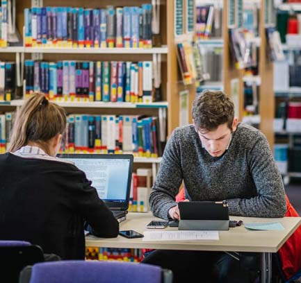 Two students studying at opposite sides of a table in a library.