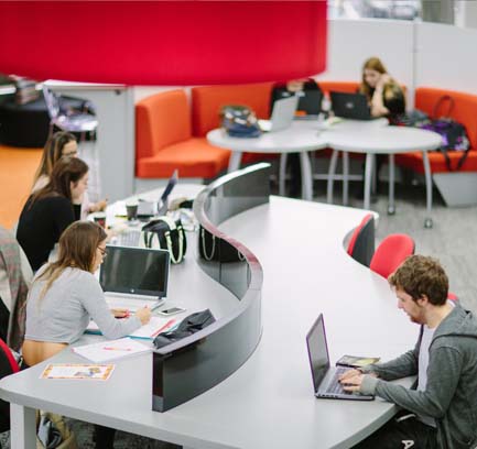 Several students working on laptops at a long, s-shaped table in a library.