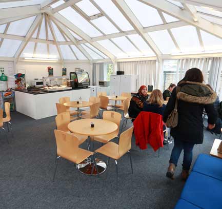 A small canteen housed inside a conservatory building with several wooden tables and chairs spaced out.