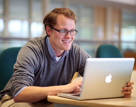 A male in smart attire, wearing glasses, sits at a desk and uses laptop.
