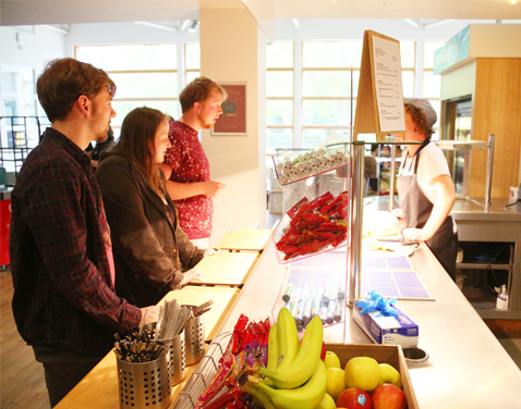 Three students in a canteen, talking to a member of kitchen staff who is standing behind a counter wearing an apron.