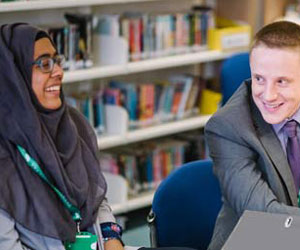 Mid-shot of two seated people in business attire, smiling at each other in a classroom setting