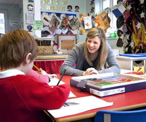 A primary school teacher in a classroom setting, smiling encouragingly while her pupil hand draws a picture