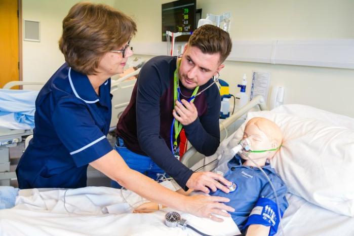 Nursing Children, A male wearing casual clothes hold a stethoscope to a child mannequin's chest. A female lecturer wearing a nurses tunic supports the male and has a hand on the mannequin's chest.