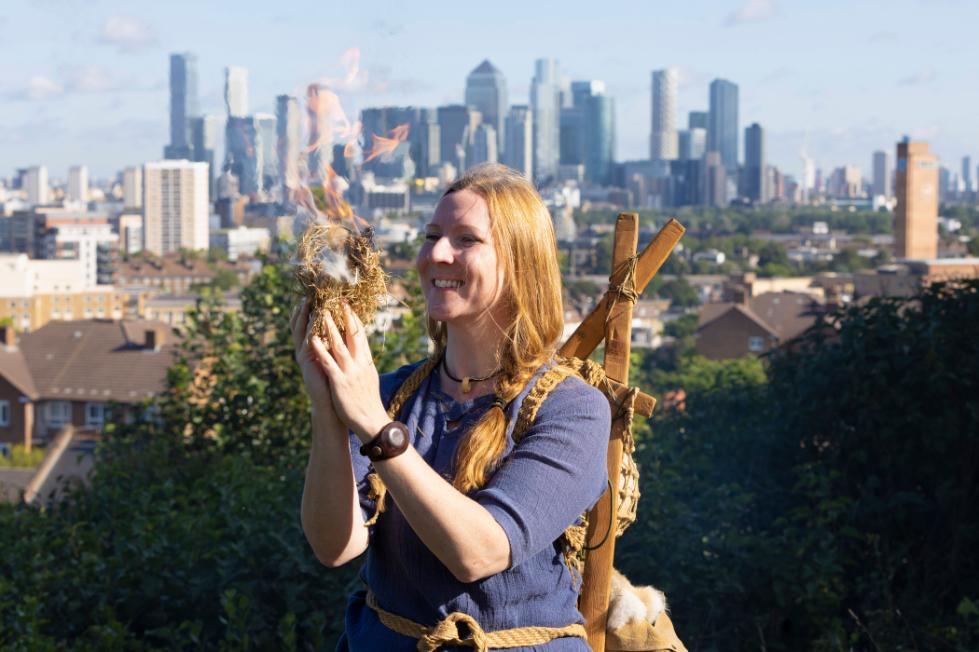 A woman stands with a city behind her burning wood and paper.